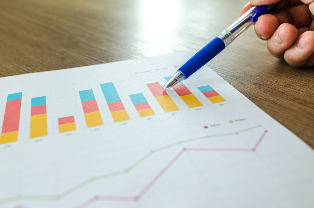 person-holding-pen-pointing-at-graph-590020 Detailed close-up of a hand pointing at colorful charts with a blue pen on wooden surface.