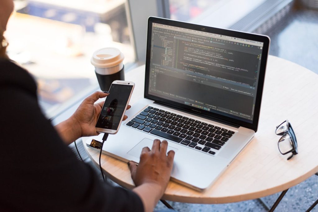 person-holding-smartphone-while-using-laptop-1181244 Developer working remotely, coding on a laptop with phone in hand, showcasing modern work culture.